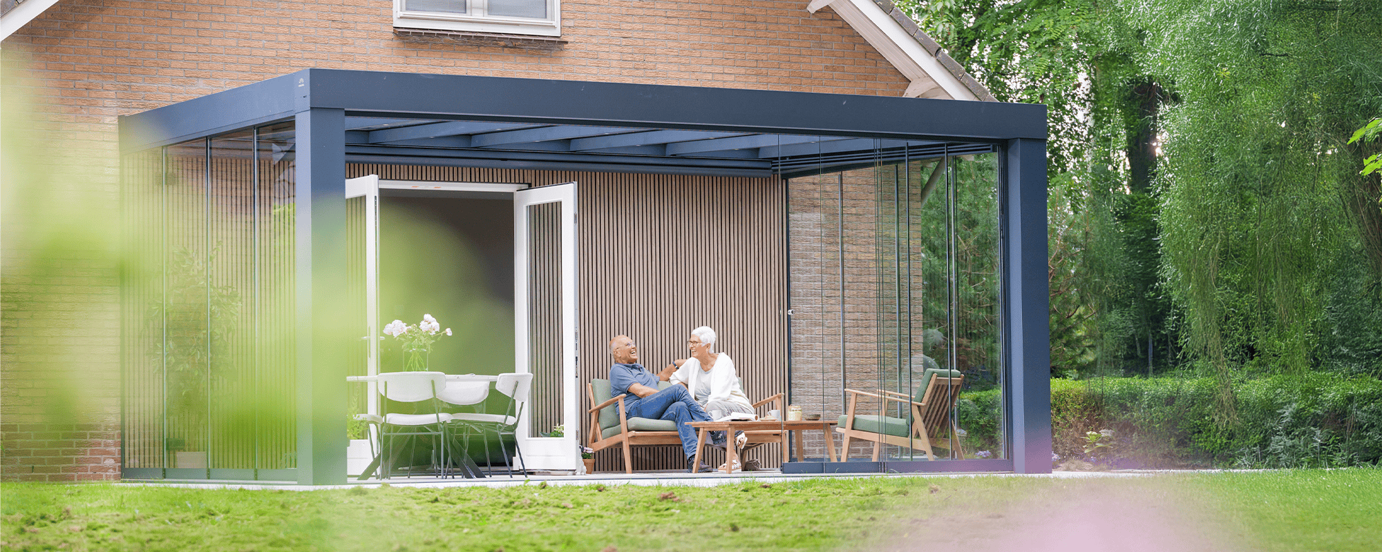 Tuinkamer met plat dak en glazen schuifwanden, aangebouwd aan een woning, sfeervol ingericht met zitmeubels waar een ouder stel samen geniet.